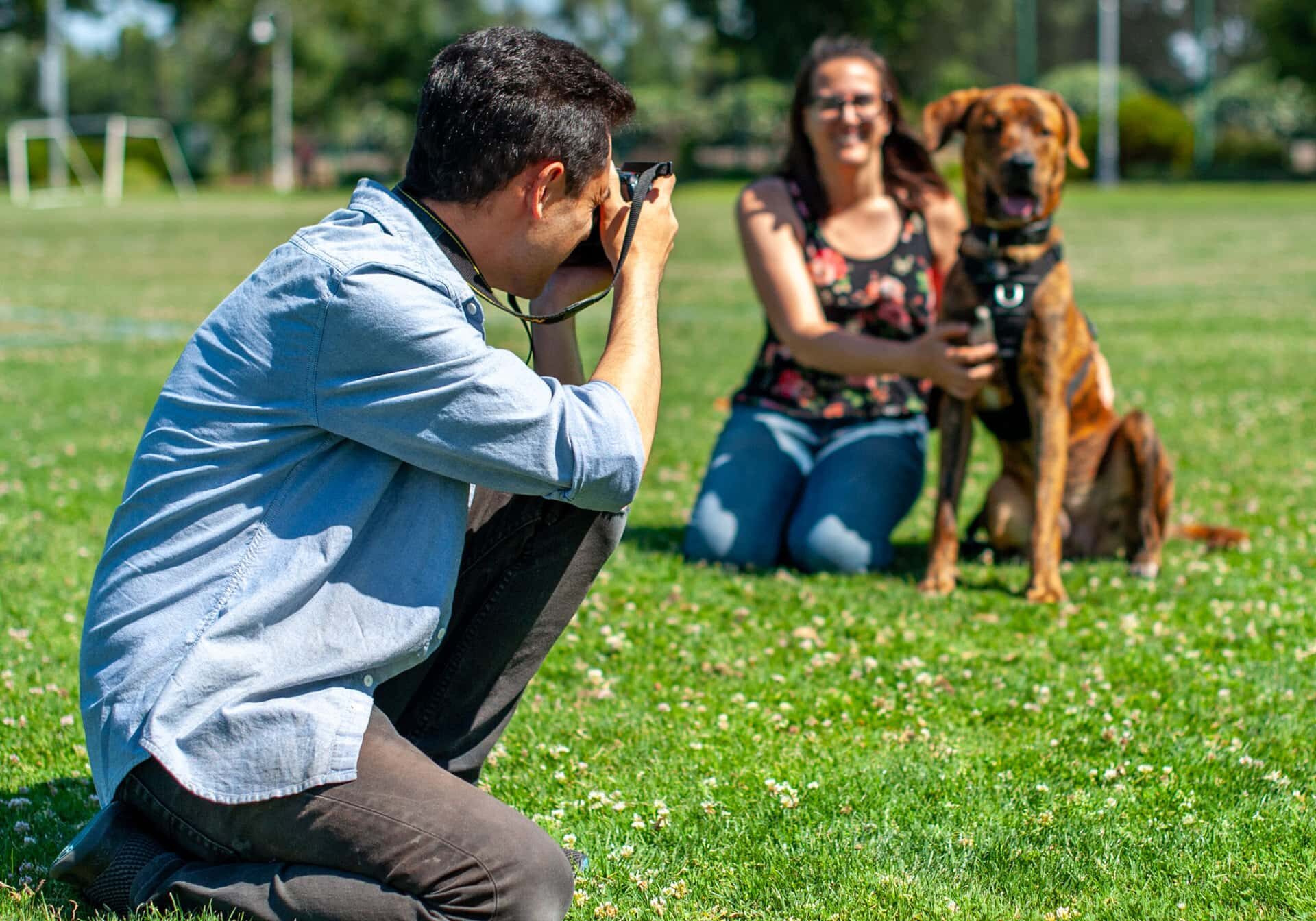 A photographer squats down while photographing a young woman posing with her dog in a park